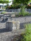 Öffentlicher Kiesplatz mit Tisch und Bänken aus Holz, Feuerschale, Granitblock und blühenden Stauden